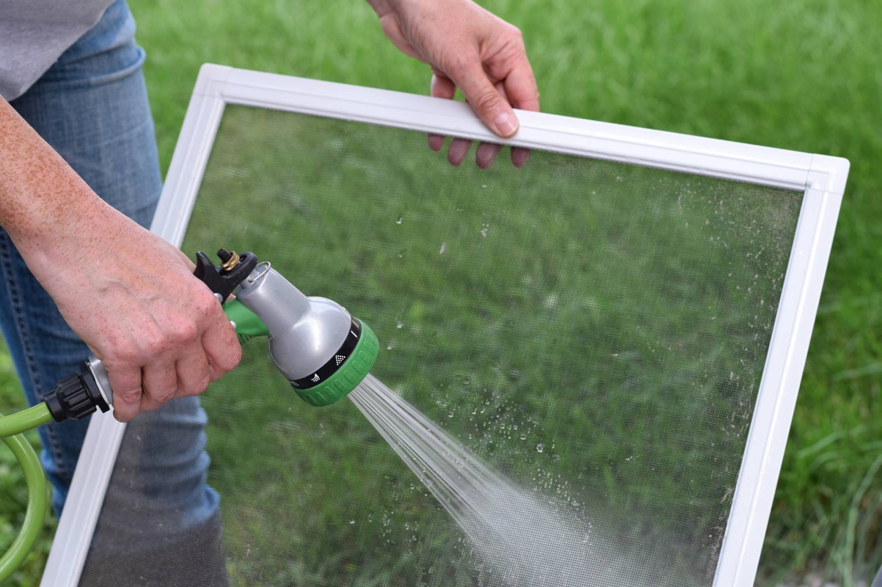 Person wearing jeans holding their removed window screen, cleaning it with a garden hose at Eagle Harbor Window Coverings near Seattle, WA